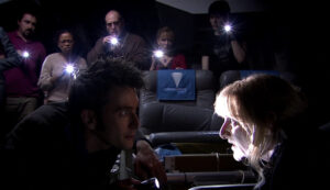 Shows a still from the Doctor Who episode called 'Midnight'. The Doctor (played by David Tennant) facing a woman called Sky (played by Leslie Sharp) her face is brightly lit from underneath. The cabin of the spaceship is dark, the passengers in the background point torches towards us.