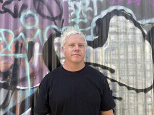 Damo Cox in black shirt standing by graffiti wall 